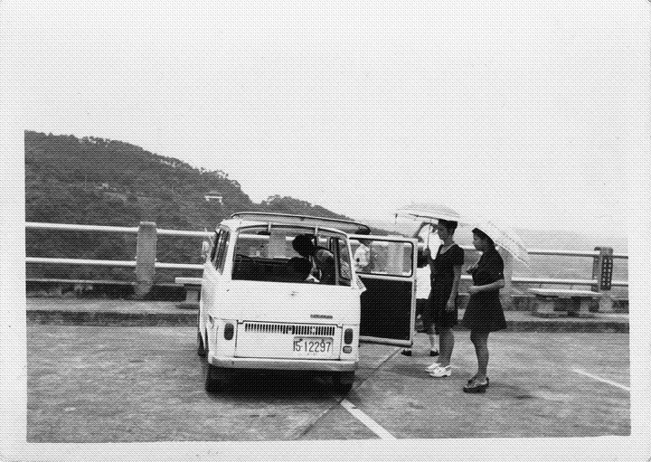 Two women holding umbrellas watching someone pack a minivan